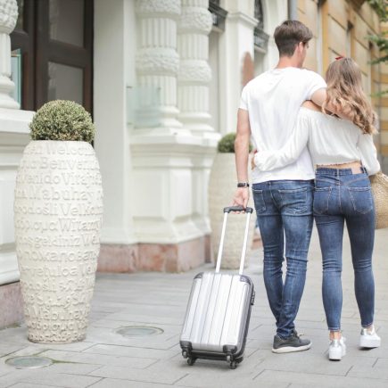 Back view of traveling couple in love wearing casual clothes walking with luggage and hugging while strolling along sidewalk together during vacation